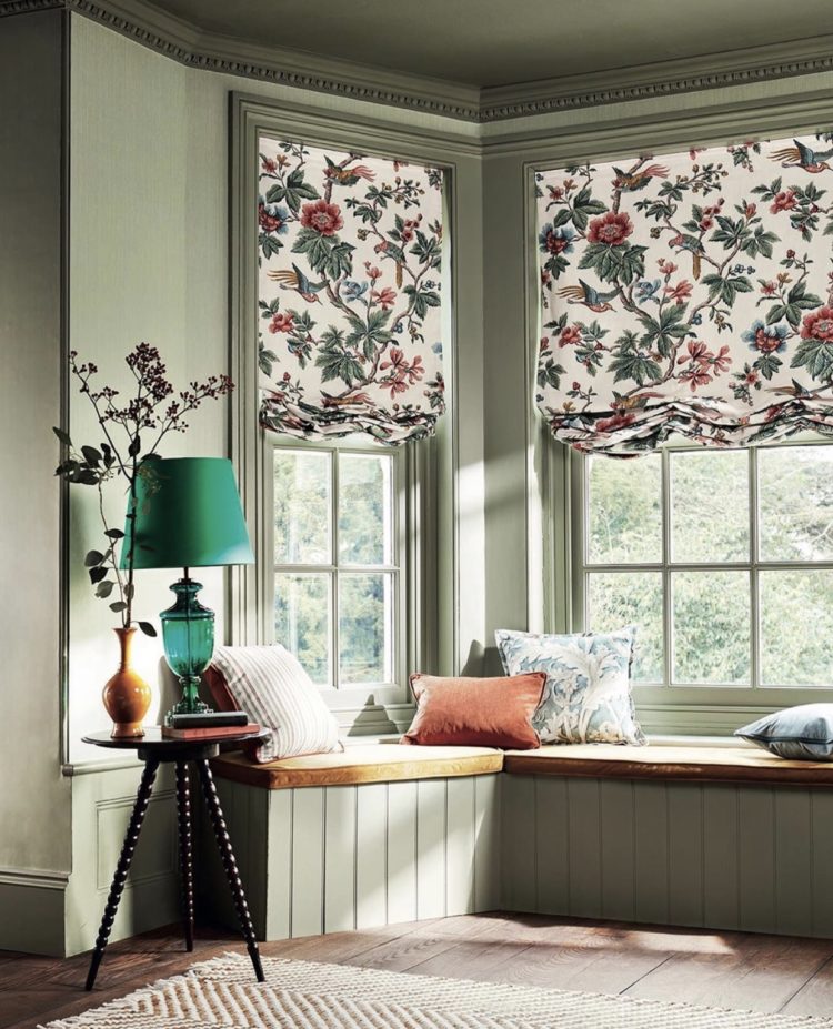 Sage green walls and woodwork around a bay window with window seat and floral roman blinds. image via sanderson
