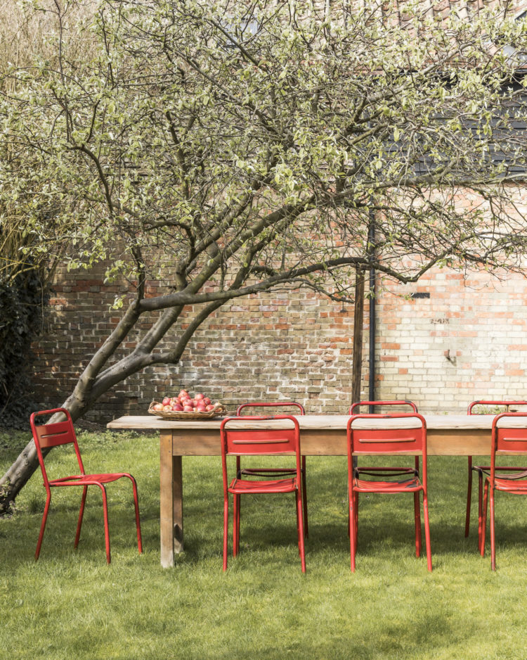 Red dining chairs, large wooden table in walled garden. Merchant & Found table with 1950s vintage original French Olix cafe chairs £675 for six